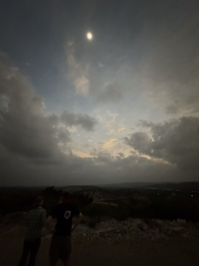 Sky and earth during total eclipse; the Sun is visible at the top of the picture, with Venus at 4pm relative. At the bottom, it is very dark; you can barely see two people a few feet away, and there are some lights from the airstrip runway in the far distance.