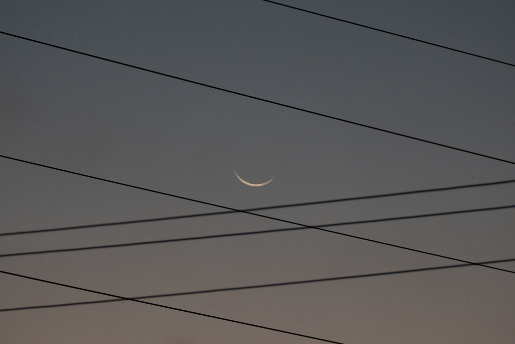 waning crescent moon seen through intersecting telephone lines in a dimly lit sky